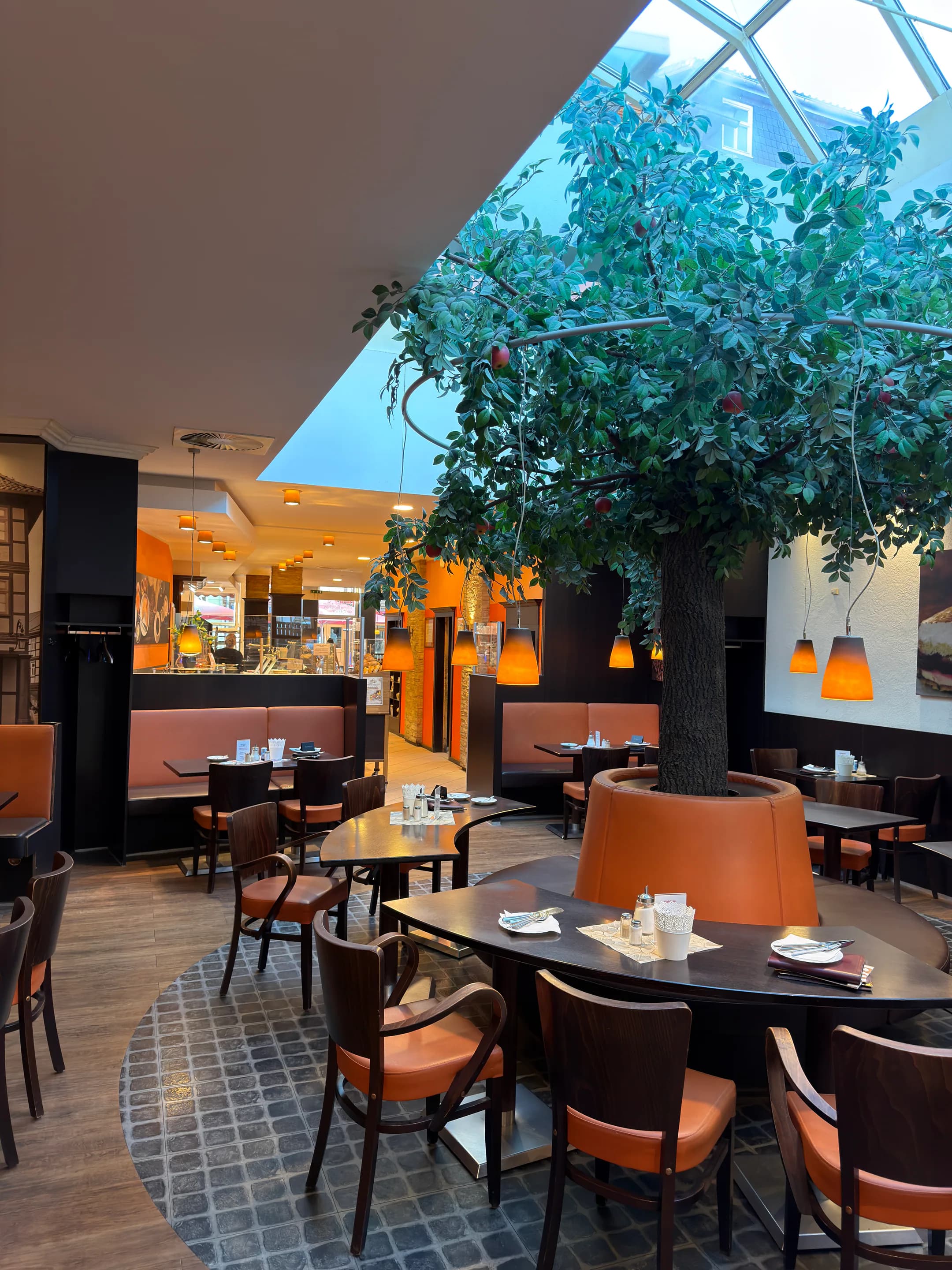 Hochformatiger Blick auf den großen Baum, die Sitzecken und orangefarbenen Pendelleuchten im Schlemmercafé