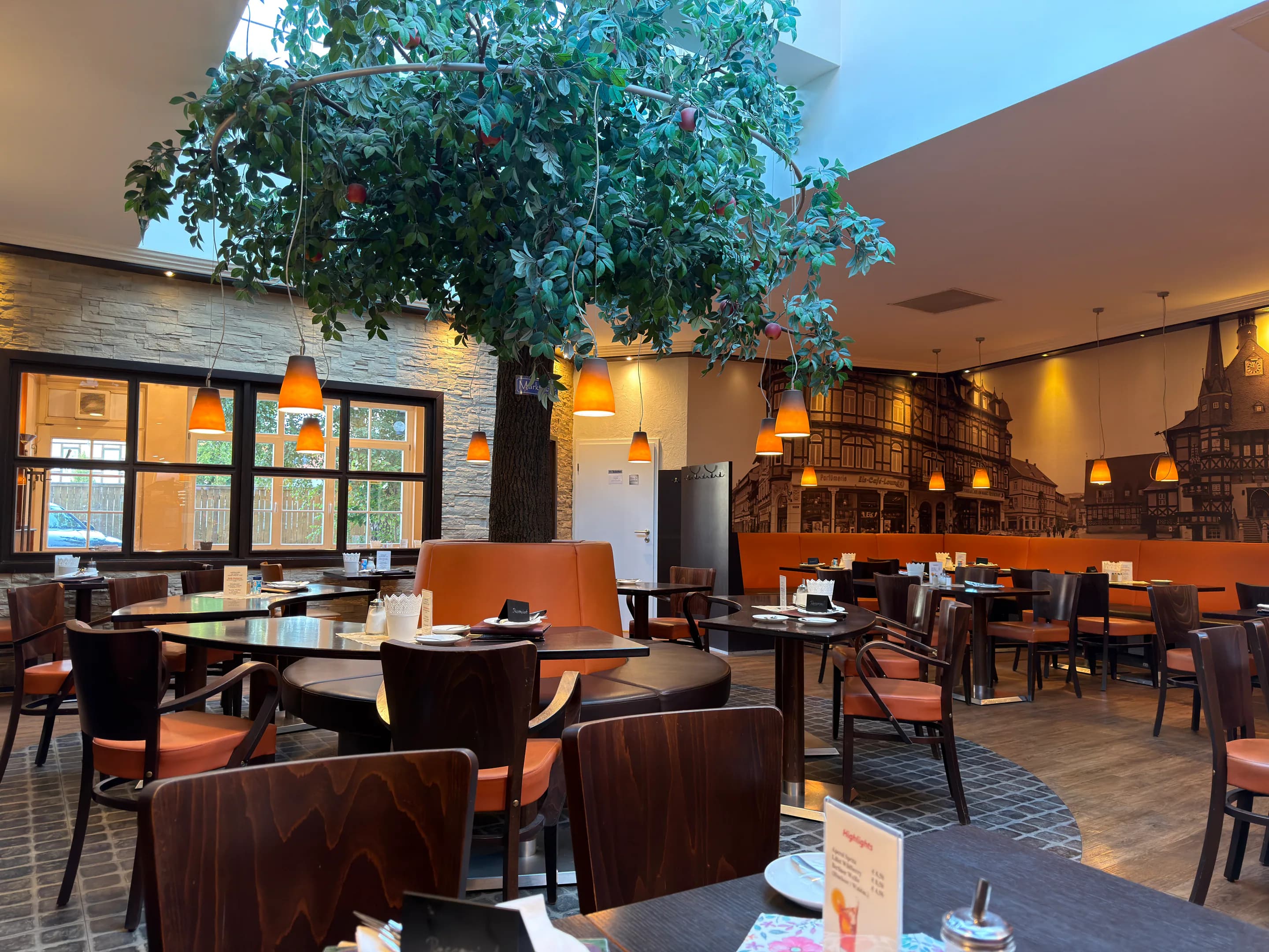 Weiter Gastraum des Schlemmercafés mit großem Baum, orangefarbenen Leuchten und gedeckten Tischen
